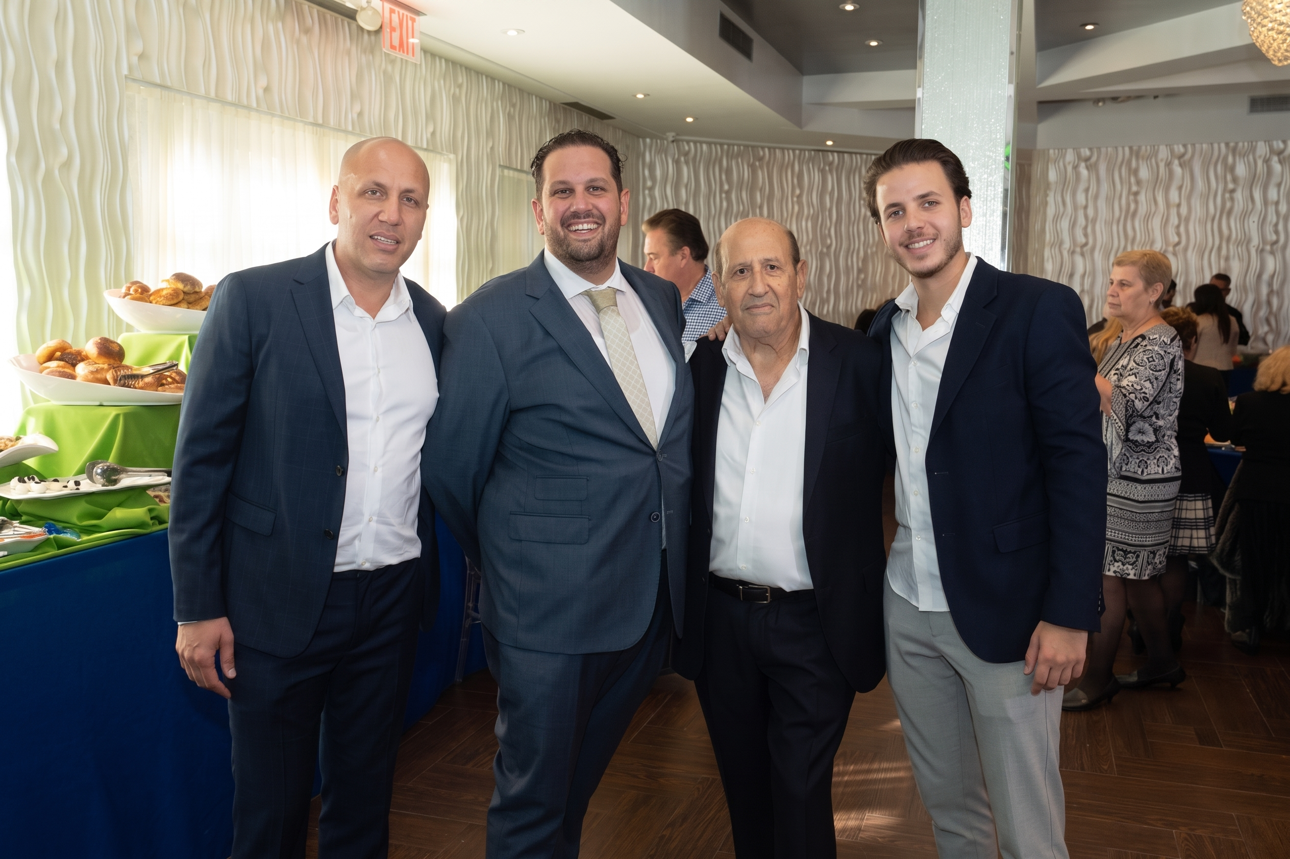 A group of four professionals in business attire pose during a catered event. Background features include a buffet with baked goods and a modern wavy-textured wall, highlighting the high-quality kitchen design services available.